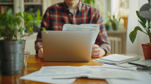 man met de papieren in de hand, achter de laptop